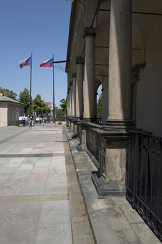 Colonnade with classical columns casting shadows on a stone pathway, flags visible in background under clear sky.