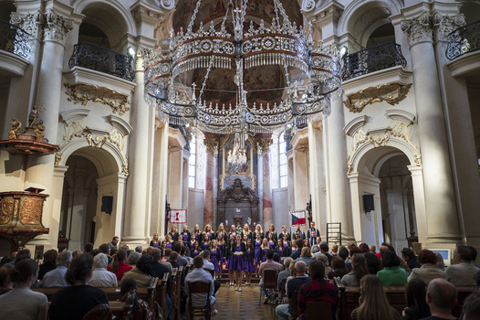 Ornate Baroque church interior with a large chandelier and choir performing on stage.