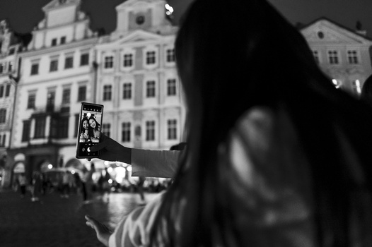 Woman holding smartphone capturing a nighttime crowd in a historic city square with classical architecture.