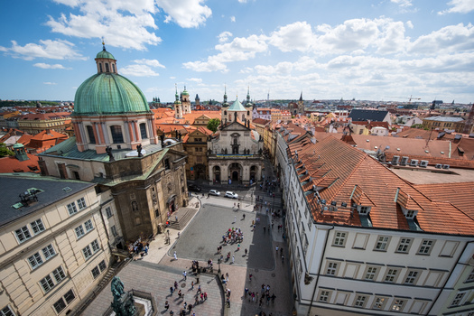 Elevated view of a European city square with terracotta roofs, a domed church, and historic architecture under blue sky with clouds.
