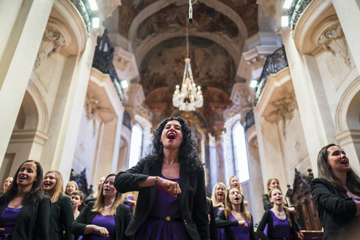 Women in purple dresses singing in a grand cathedral with ornate ceiling and chandelier.