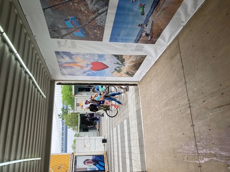 Outdoor exhibition space with large printed photographs displayed on white panels under a metal roof structure.