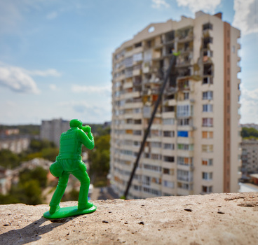 Green toy soldier figurine in sharp focus on concrete ledge, with blurred apartment building and sky behind.