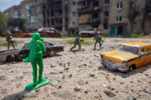 Green toy soldier figure in foreground with miniature vehicles and toy soldiers arranged on sandy ground before damaged apartment building.