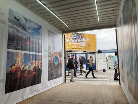 Interior of a covered exhibition space with large-format photographs on white panels and visitors walking through.