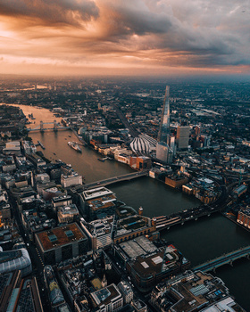 Aerial view of a sprawling city with a meandering river, bridges, and dense urban development under dramatic sunset clouds.