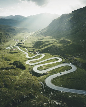 Winding mountain road cutting through lush green valley with dramatic sunlit ridges.