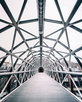 Symmetrical view down a modern pedestrian bridge with geometric steel truss framework overhead.