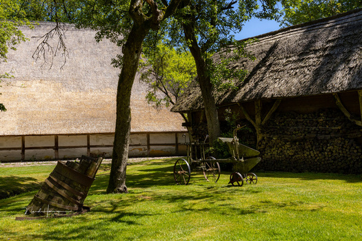 Rustic farmyard with thatched barn, wooden fence, vintage farm equipment, and lush green lawn beneath mature trees.