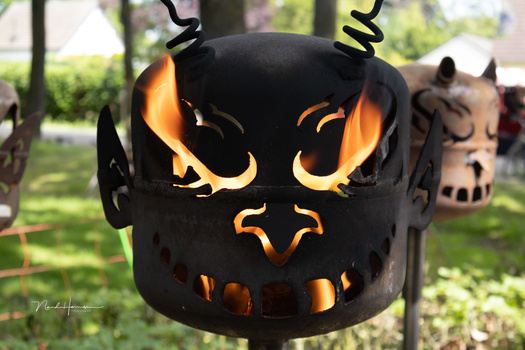 Black metal fire pit with carved demon face glowing with orange flames at dusk in outdoor garden setting.