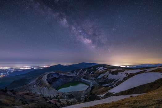 Milky Way arching over volcanic crater lakes with snow patches and distant city lights on horizon.