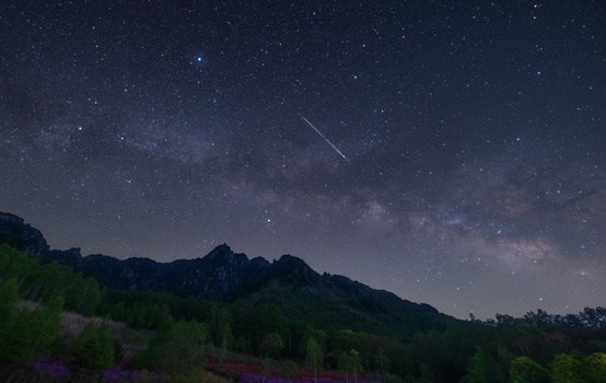 Milky Way galaxy stretching across night sky above silhouetted mountains with a meteor streak visible.