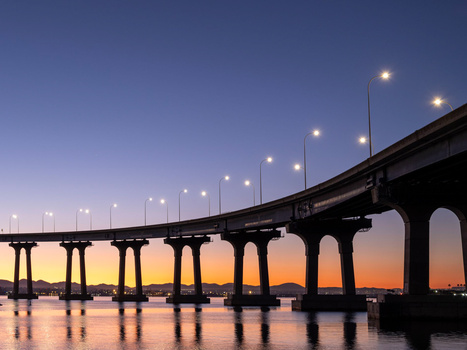 Illuminated bridge spanning across calm water at twilight with warm orange and blue sky.