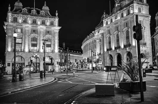 Nighttime urban plaza with illuminated neoclassical buildings and wet pavement reflecting lights.