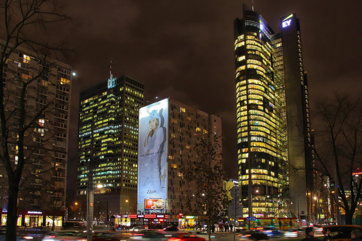 Nighttime urban street scene with illuminated skyscrapers and a large vertical advertisement display.