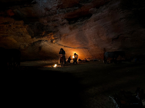 Group of people gathered around warm candlelight inside a dark cave illuminated by golden firelight.