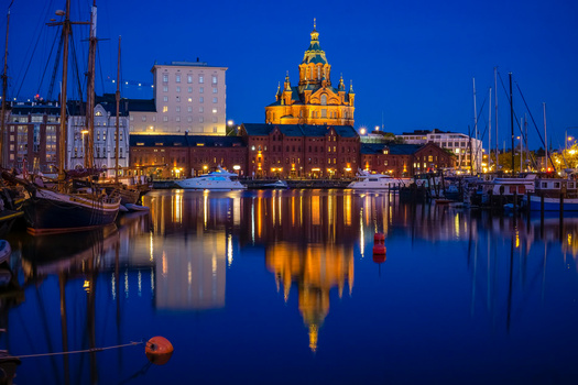 Illuminated cathedral with golden dome reflected in still harbor water at twilight, surrounded by sailing boats.