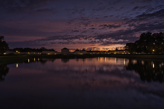 Twilight landscape with calm water reflecting illuminated buildings and trees along the shoreline.