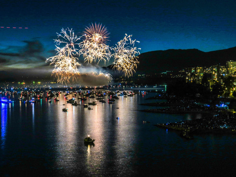Fireworks burst over a crowded bay at twilight, reflecting in calm water below mountains.