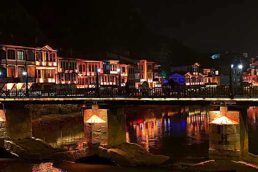 Riverside buildings illuminated in warm orange and red light reflected in dark water below a stone bridge at night.