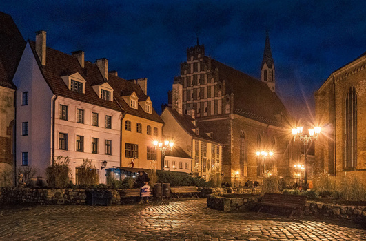Historic European town square at dusk with illuminated medieval buildings and church spire.