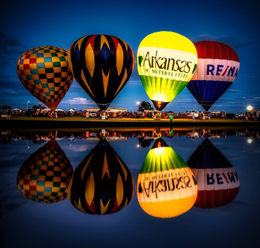 Four illuminated hot air balloons reflected in still water at dusk with a bridge and lights in the background.