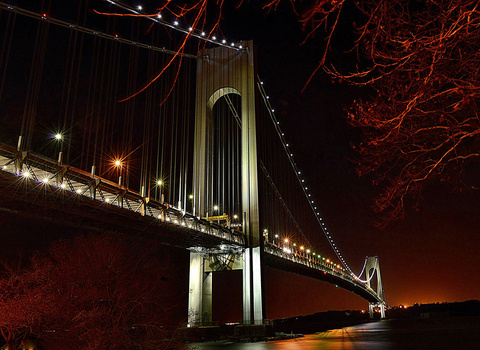 Suspension bridge illuminated at night with warm golden lights reflecting on dark water below.