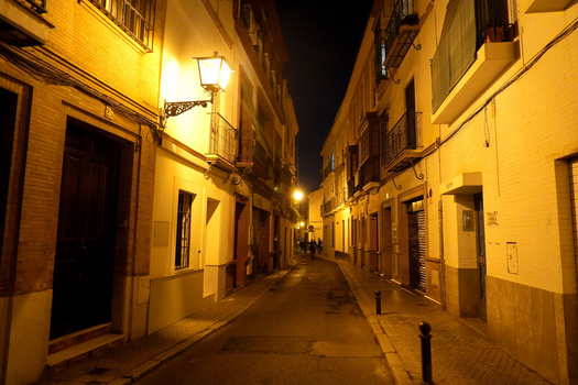 Narrow European street at night illuminated by warm streetlights, with yellow-toned buildings lining both sides.