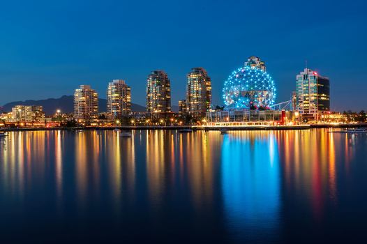 Illuminated city skyline reflected in calm water at dusk, with a glowing spherical structure prominently featured.