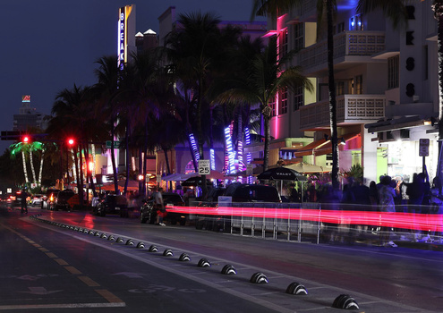 Neon-lit street at dusk with palm trees, colorful building signs, and light trails from passing vehicles.