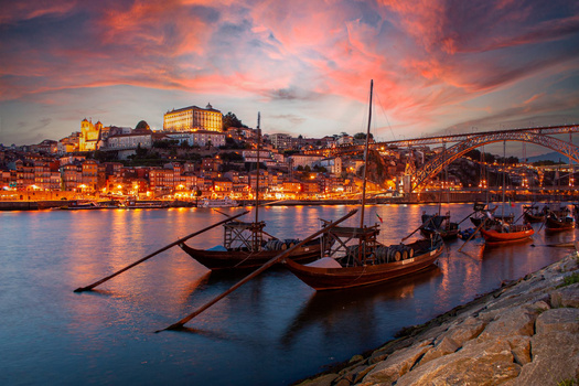 Traditional wooden boats moored in a river at dusk, with illuminated hillside buildings and bridge in background.