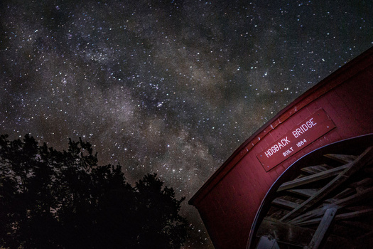Milky Way galaxy stretching across night sky above silhouetted trees and a red bridge structure.