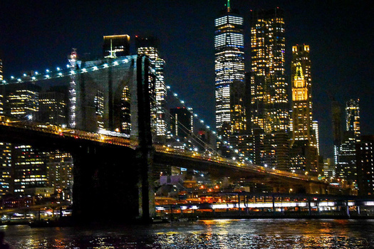 Brooklyn Bridge spanning the East River at night, illuminated by warm golden lights with Manhattan skyscrapers glowing in the background.