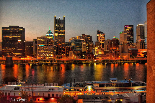 City skyline at dusk reflected in calm water, with illuminated buildings and a riverboat in the foreground.