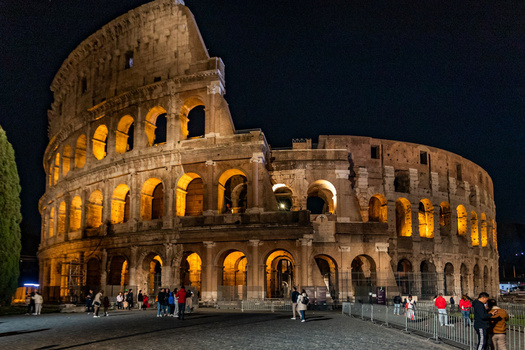 Ancient Roman amphitheater illuminated at night with golden lighting highlighting its multi-tiered arches and weathered stone facade.