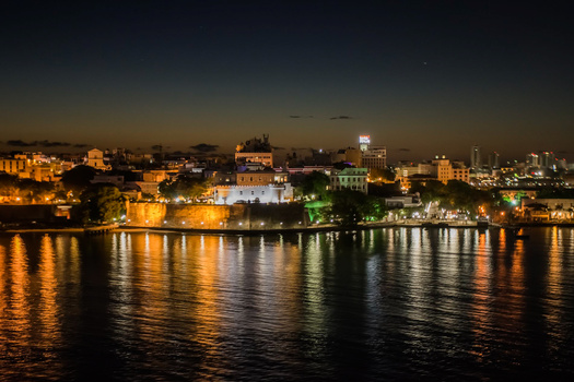 Waterfront cityscape at dusk with illuminated buildings reflected in calm water.