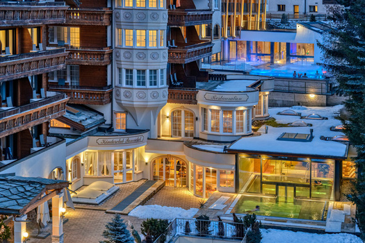 Luxury resort courtyard at dusk with illuminated buildings, pool, and snow-covered rooflines.