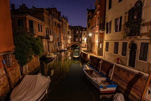 Venetian canal at twilight with moored gondolas and illuminated historic buildings.