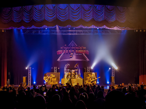 Concert stage with dramatic blue and gold lighting, ornate set design, and large crowd silhouettes in foreground.