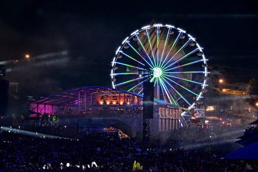 Illuminated Ferris wheel and colorful stage structures at a nighttime festival or event with crowds below.