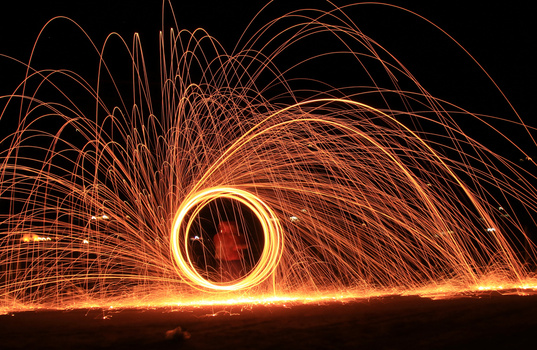 Steel wool spinning creates concentric circles of glowing orange and yellow light trails against a dark night sky.
