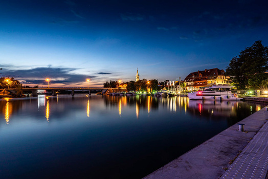 Riverside town at dusk with illuminated buildings and bridge reflecting in calm water.