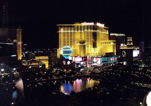 Nighttime cityscape of illuminated resort buildings reflected in water, with colorful neon signage visible along the waterfront.