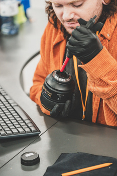Photographer in orange jacket examining a digital camera while seated at a desk with laptop and workspace.