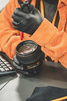 Photographer in orange jacket holding a Nikon lens with black gloved hands over a workspace.