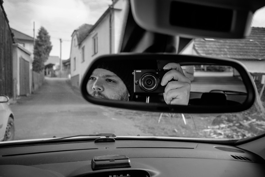 Photographer's reflection in car mirror holding vintage rangefinder camera, captured in black and white.