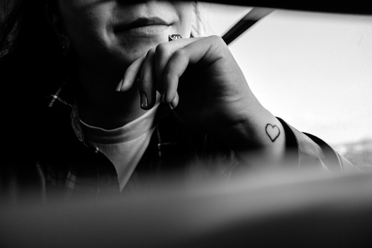 Black and white portrait of person with heart tattoo on shoulder, hand near face in contemplative pose.