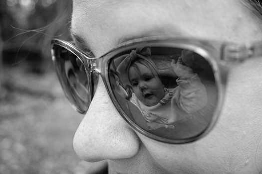 Close-up of a person wearing large clear-framed glasses with a reflection of a child visible in the lens.