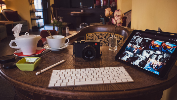 Flat lay of a photographer's workspace with a vintage camera, keyboard, tablet displaying photos, coffee cups, and decorative elements on a round wooden table.