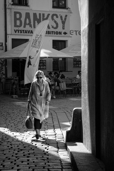 Woman walking through a narrow European-style alley with strong directional sunlight creating geometric shadows on cobblestone pavement.